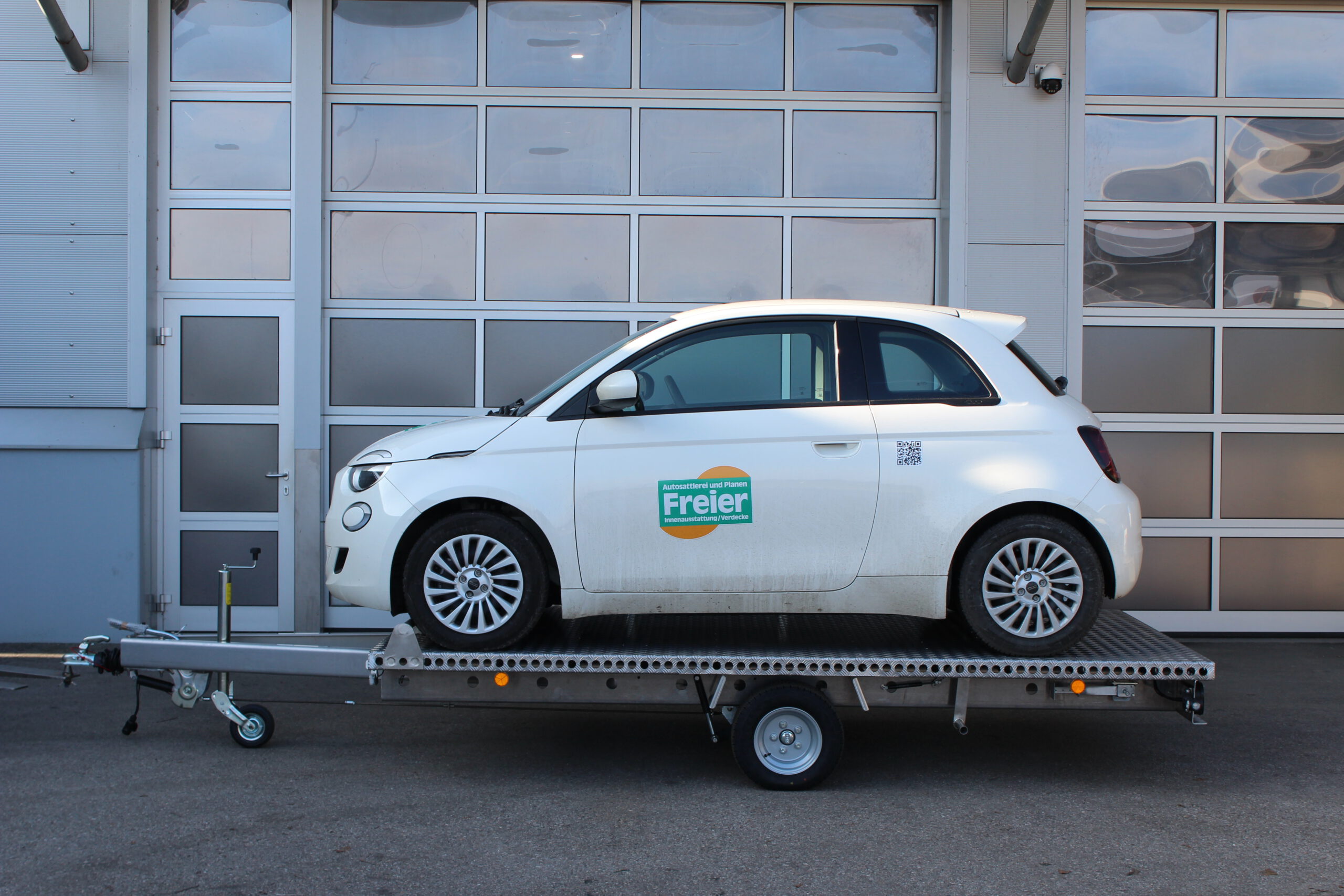 Aluminium Anhänger Autotransporter, Hochlader Plattform beladen mit einem weißen Fiat 500E vor einem Hallentor