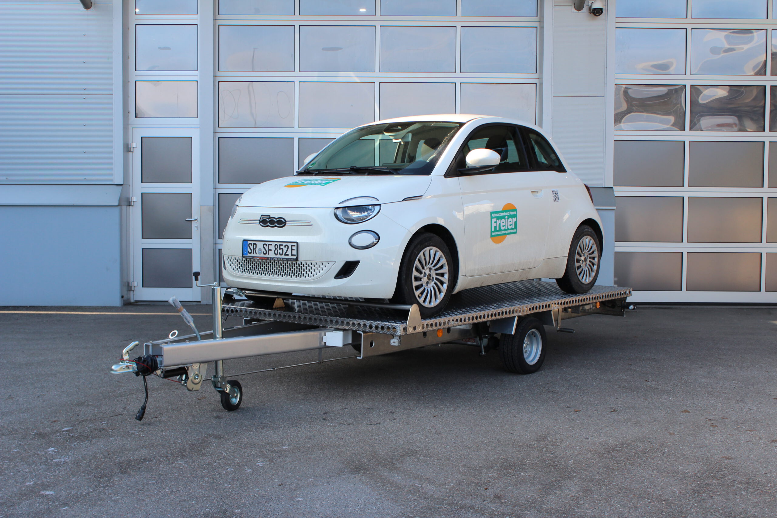 Aluminium Anhänger Autotransporter, Hochlader Plattform beladen mit einem weißen Fiat 500E vor einem Hallentor